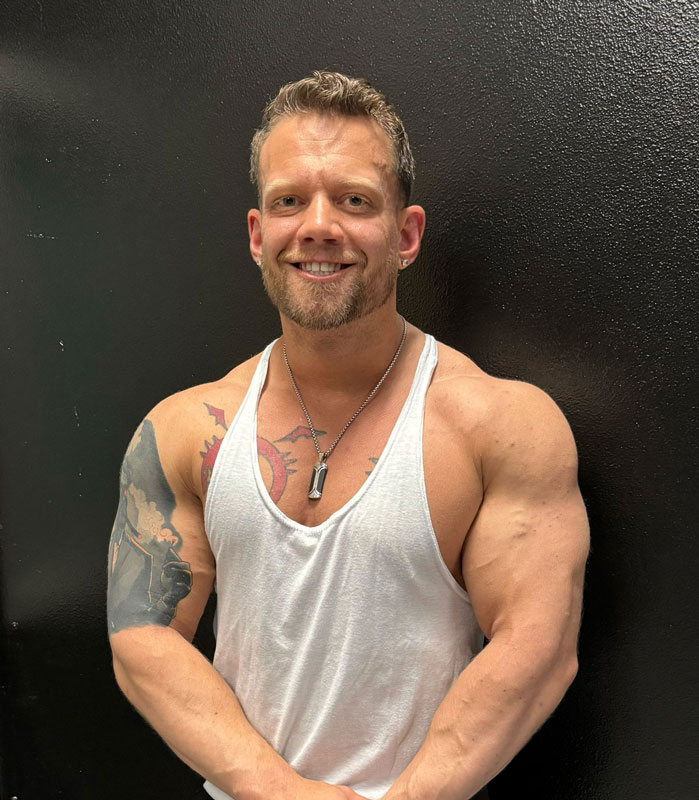 A photo of a coach smiling & flexing his muscles in a white tank top.