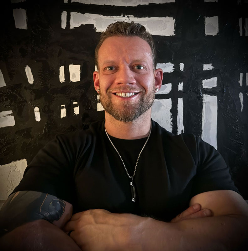 A photo of a coach smiling with his arms crossed in a black t-shirt.